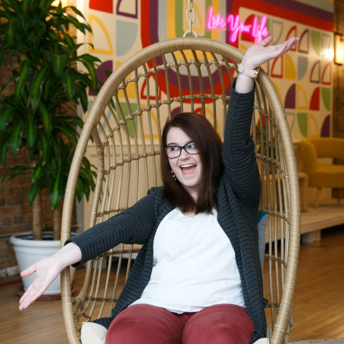 Gabby smiling widely in a hanging bamboo chair inside of a modern office with arms and legs wide