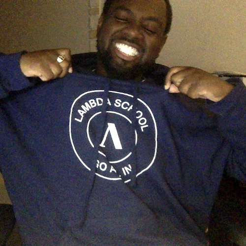 Junior with a wide smile drawing attention to his Lambda School sweatshirt he proudly wears.